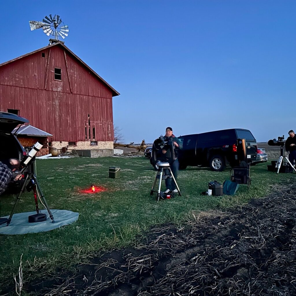 Members setting up telescopes on the grass beside a red barn at dusk