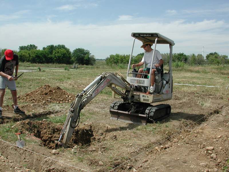 A member at the controls of a mini excavator digging a foundation trench in an open field while another member works with a pick