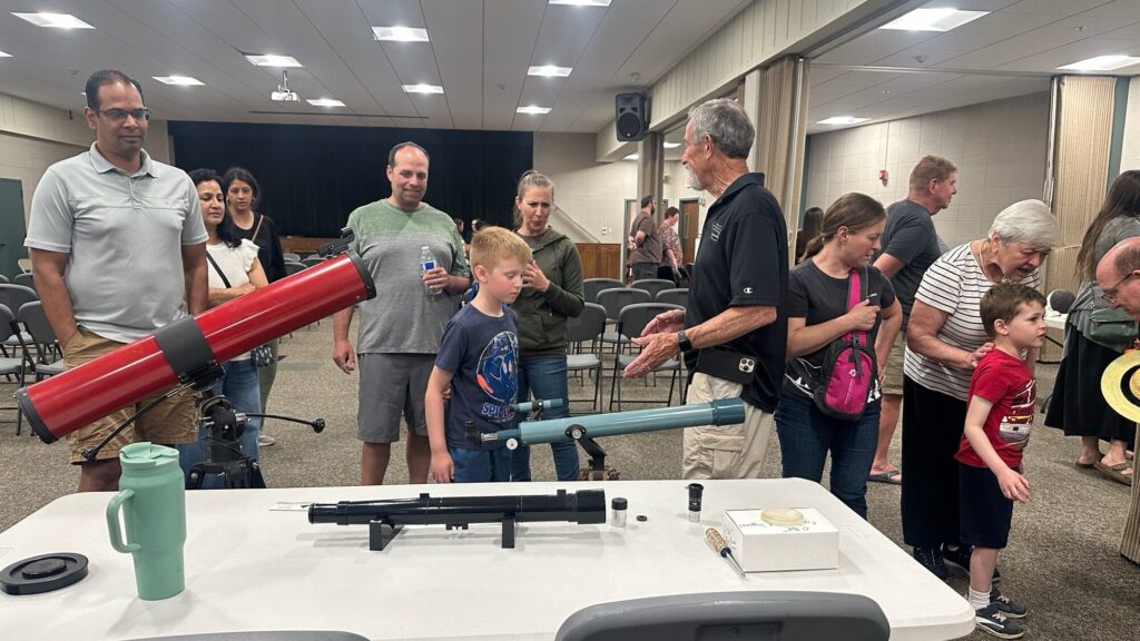 A member showing a boy and his family a row of small telescopes at an indoor event