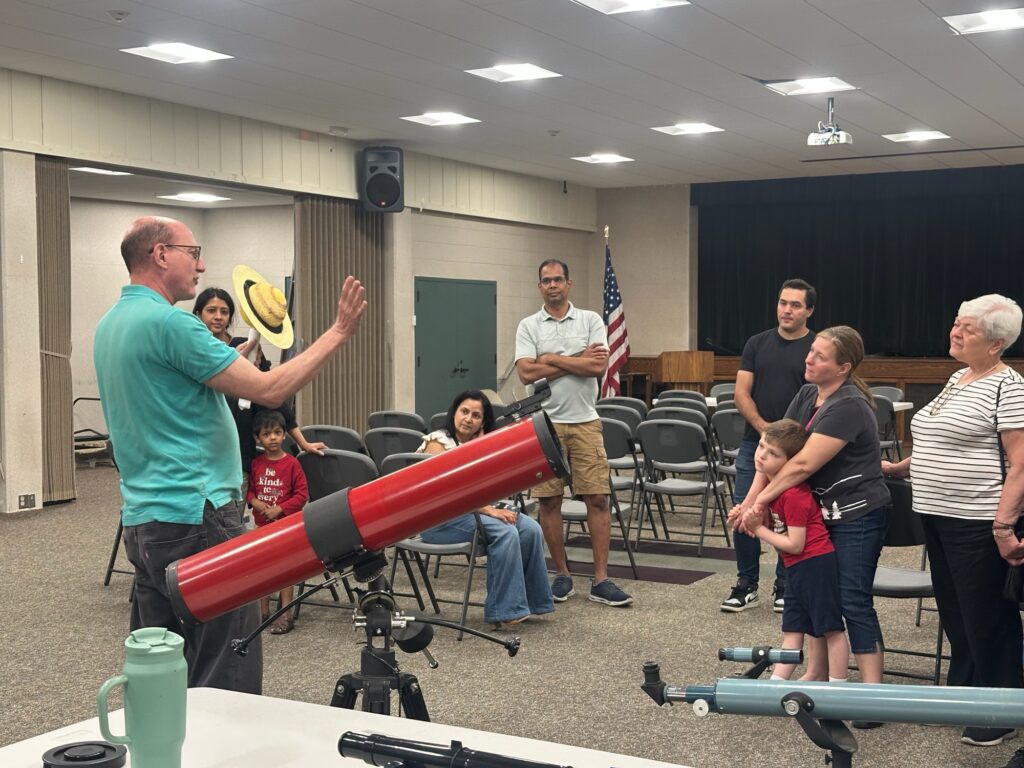 A member giving a talk beside a red telescope to families seated in a meeting room
