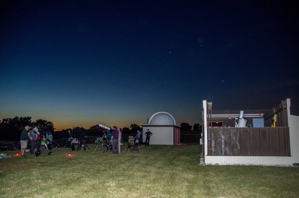 Members with telescopes on the lawn at the Astronomy Education Center at dusk, the Riley Observatory dome behind them