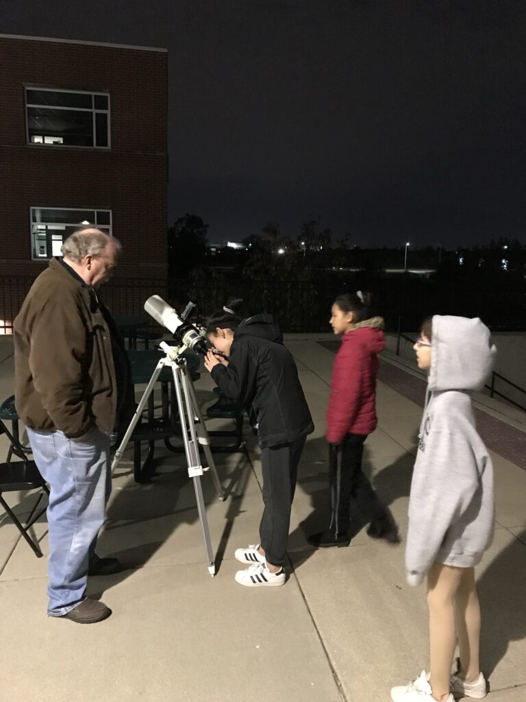 A member showing students the view through a telescope outside a school at night