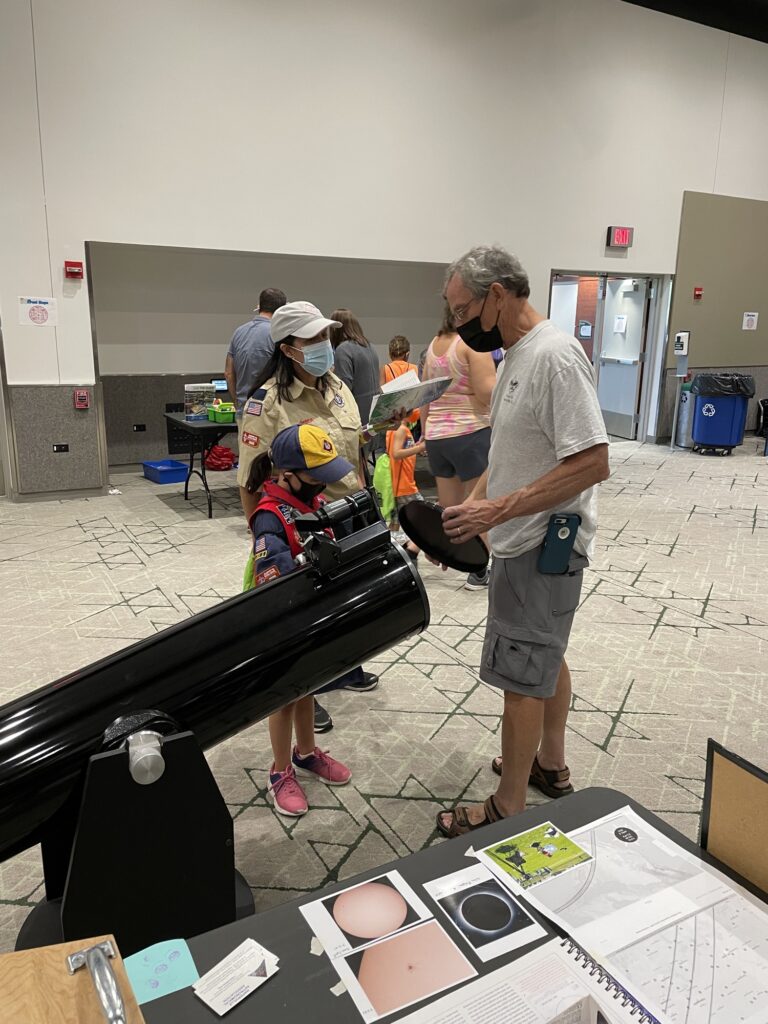 A member talking with a scout beside a large telescope at an indoor outreach event